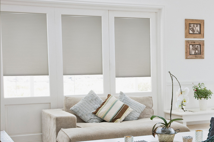 Cellular shades are energy-conservative for your living spaces. Light gray cellular shades on three separate windows in large light wood and beige colored living room.
